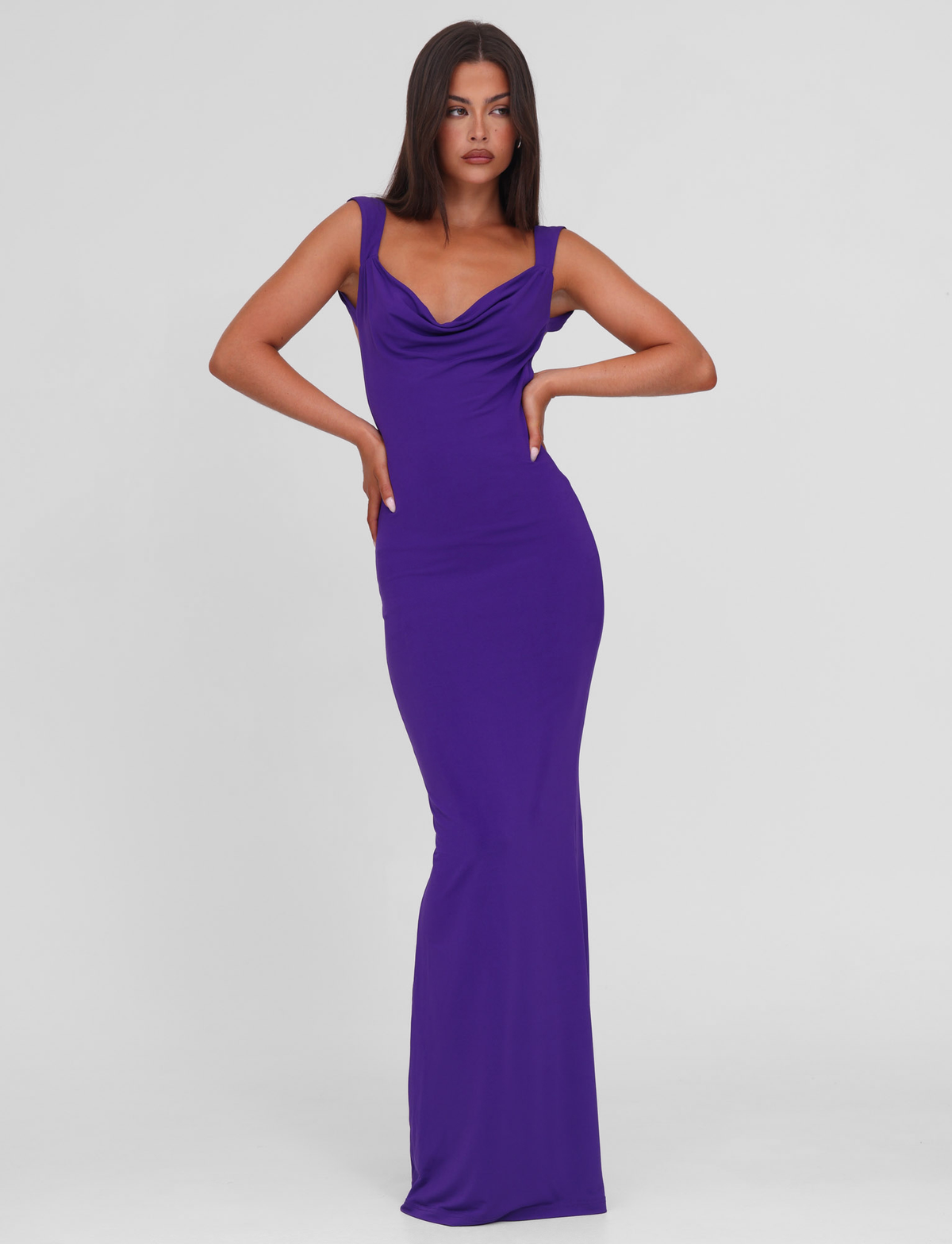 A woman stands confidently in the TIGERMIST ZITA MAXI DRESS - PURPLE, a sleeveless, floor-length bodycon dress with a draped neckline, set against a plain white background.