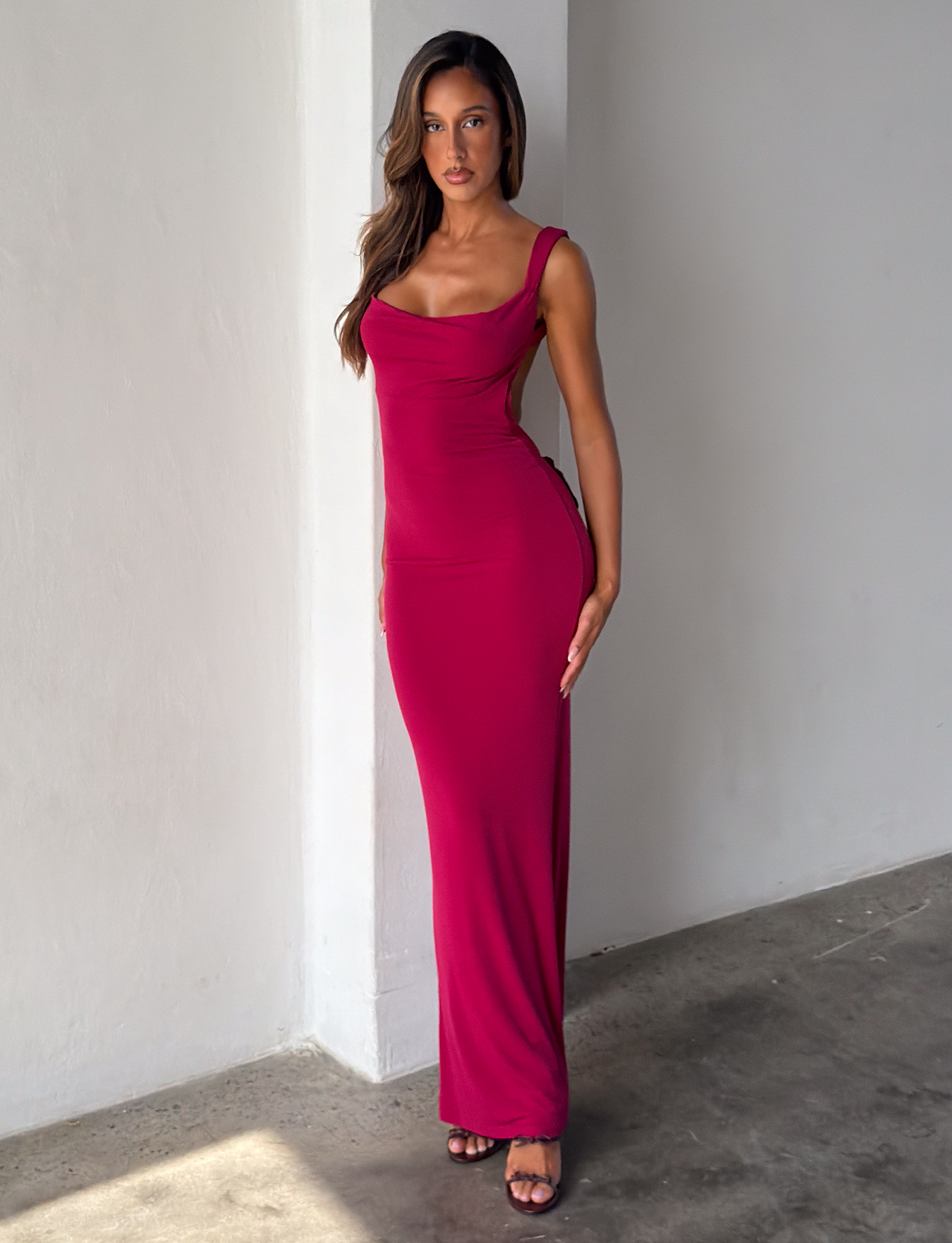 A woman stands indoors against a white wall, modeling the TIGERMIST ZITA DRESS in Deep Red (Salsa), a form-fitting, sleeveless, floor-length dress, paired with black heeled sandals. She has long wavy brown hair and a confident, relaxed expression.