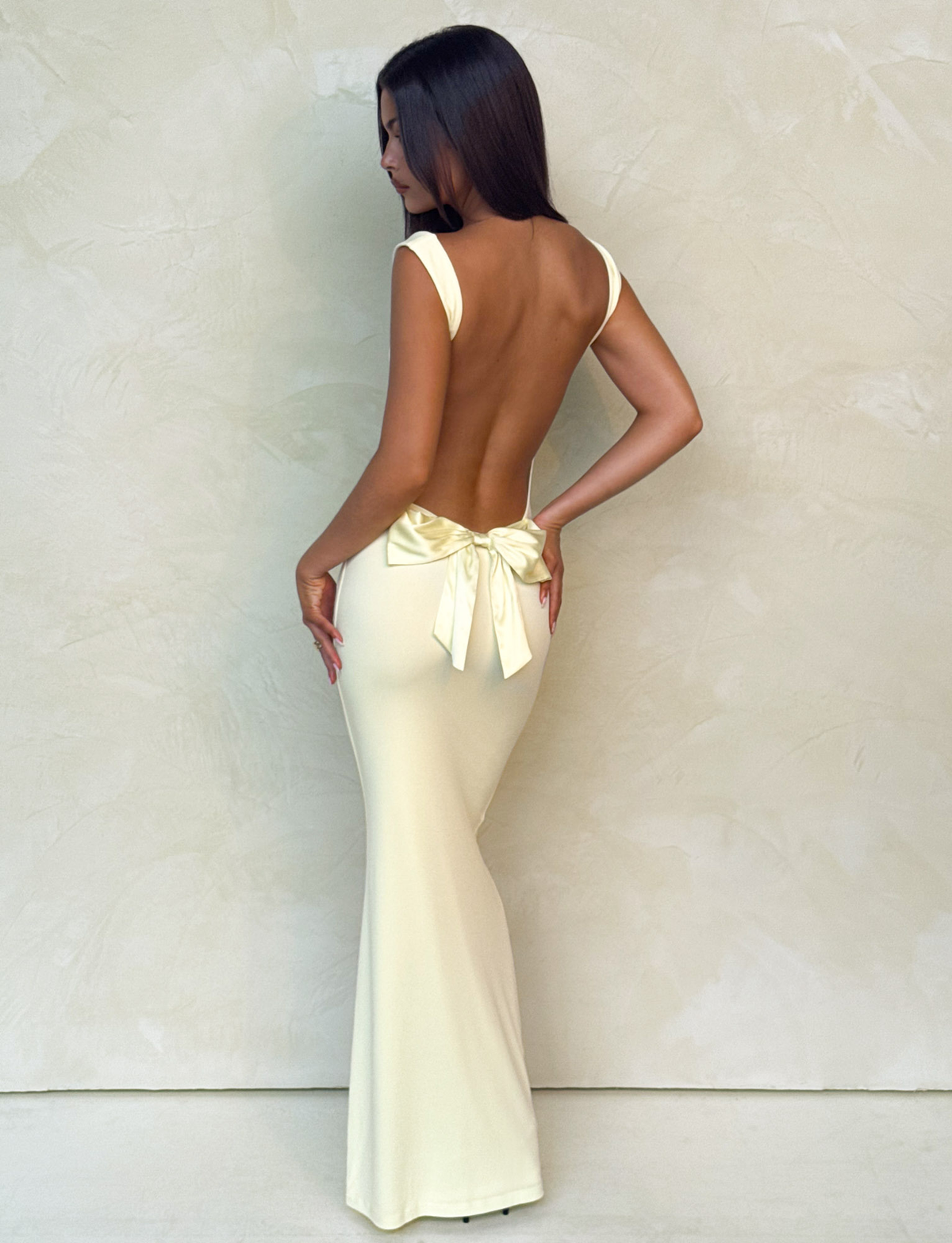 A woman with long dark hair poses in the TIGERMIST ZITA MAXI DRESS - LEMON, a pastel yellow, backless, bodycon dress with a large bow at the lower back, standing against a light textured wall.