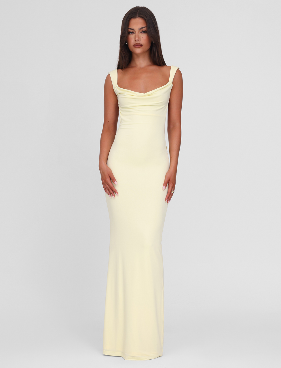 A woman with long brown hair wears the TIGERMIST ZITA MAXI DRESS - LEMON, a pastel yellow, floor-length, sleeveless gown with a draped neckline, standing against a plain light gray background.