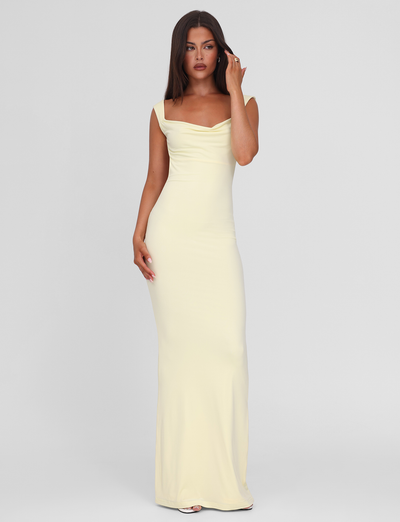 A woman with long brown hair wears the sleeveless, floor-length ZITA MAXI DRESS - LEMON by TIGERMIST, featuring a draped neckline, standing against a plain light gray background.