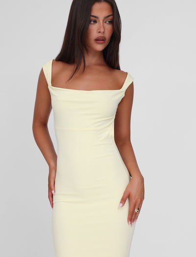 A woman with long dark hair wears the TIGERMIST ZITA MAXI DRESS - LEMON, an off-shoulder, bodycon silhouette, posing with one hand on her hip against a plain light background.