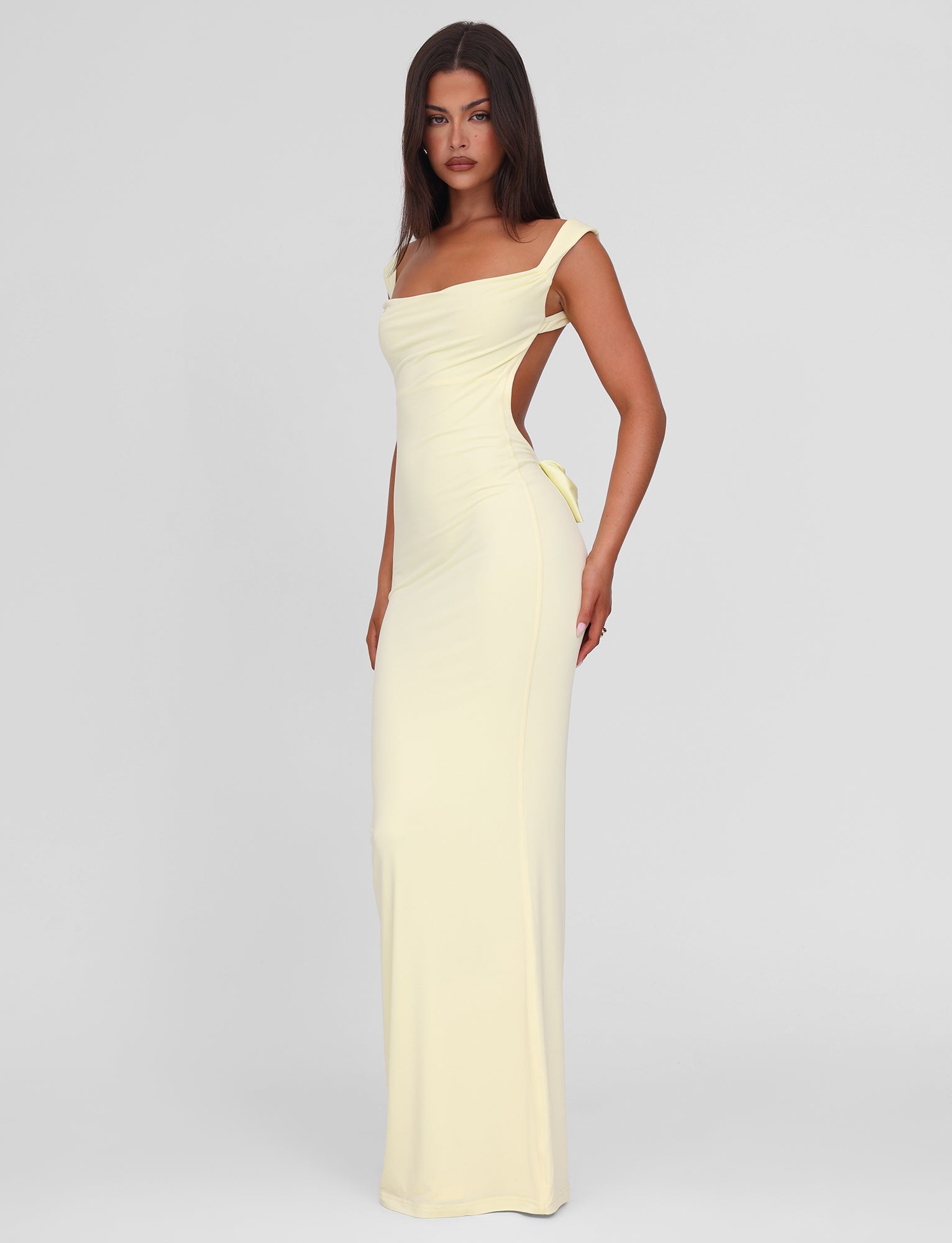 A woman poses confidently against a plain background in the TIGERMIST ZITA MAXI DRESS - LEMON, a pastel yellow, figure-hugging dress with shoulder straps and an open back. Her hair is straight and worn loose.