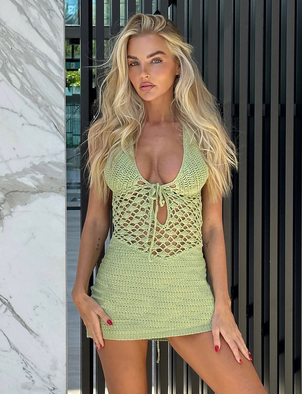 A woman with long blonde hair stands in front of a black striped background and a marble-patterned wall. She is wearing the ISABELL DRESS in pastel green from TIGERMIST. She is looking directly at the camera with a neutral expression, her hands resting on her hips.