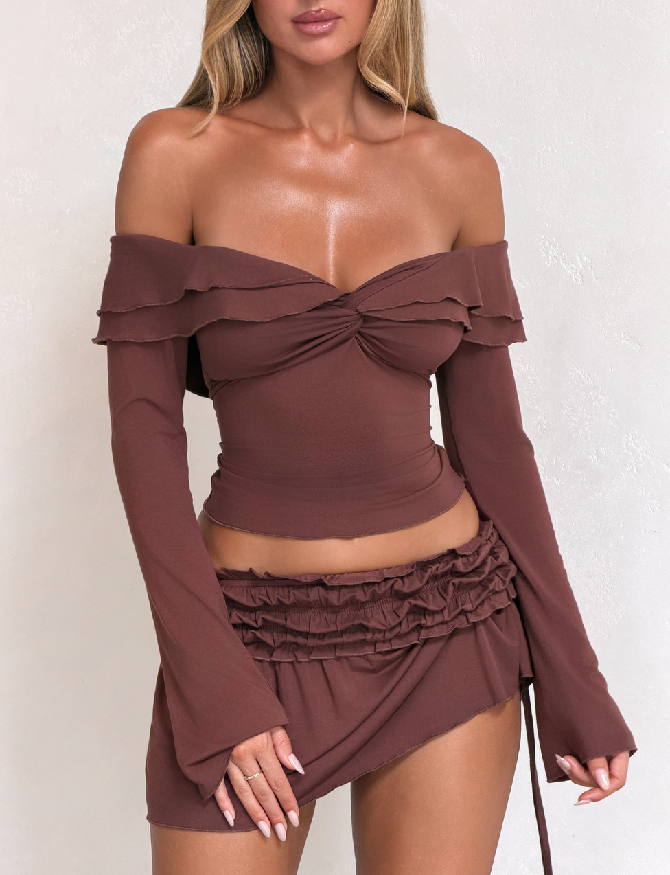 A woman models the TIGERMIST DENALI LONG SLEEVE TOP in chocolate, featuring an off-shoulder neckline and ruffle details, paired with a matching brown ruched mini skirt against a light textured background.