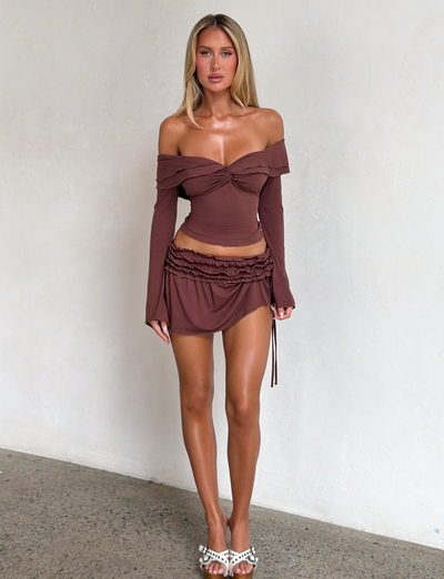 A woman with long blonde hair poses indoors against a white wall, wearing the TIGERMIST DENALI LONG SLEEVE TOP in chocolate with an off-shoulder neckline and matching skirt, styled with white heeled sandals adorned with decorative details.