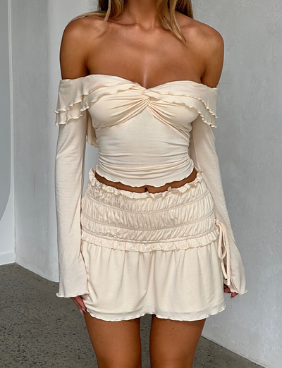 A woman wears the TIGERMIST DENALI LONG SLEEVE TOP in White: Bone, with off-the-shoulder sleeves and ruffle details, paired with a matching high-waisted ruched mini skirt against a minimalist indoor backdrop.