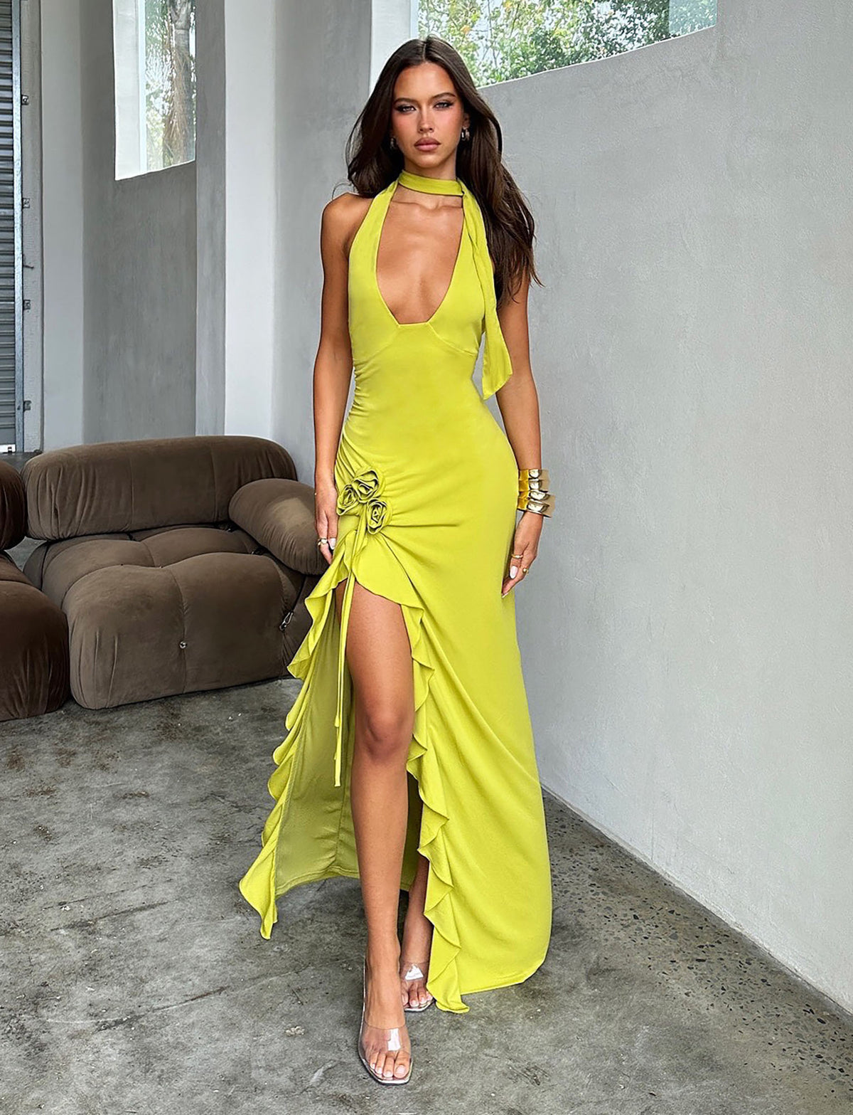 A woman stands in a modern, minimalistic room with a taupe sofa. She is wearing a bright chartreuse, sleeveless Tigermist FAORA DRESS with a high slit and ruffled edges, accessorized with a matching neck scarf and metallic bracelets. She has long, straight hair and is looking directly at the camera.