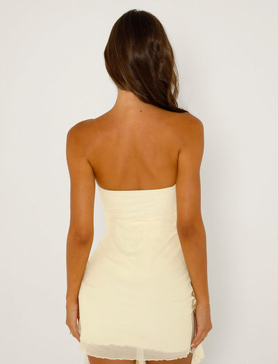 A woman with wavy brown hair stands facing away, showcasing the intricate back details of a strapless, yellow dress from Tigermist. The SADIA DRESS - YELLOW : BUTTER is fitted and falls to mid-thigh length, exuding elegance against a plain white background.