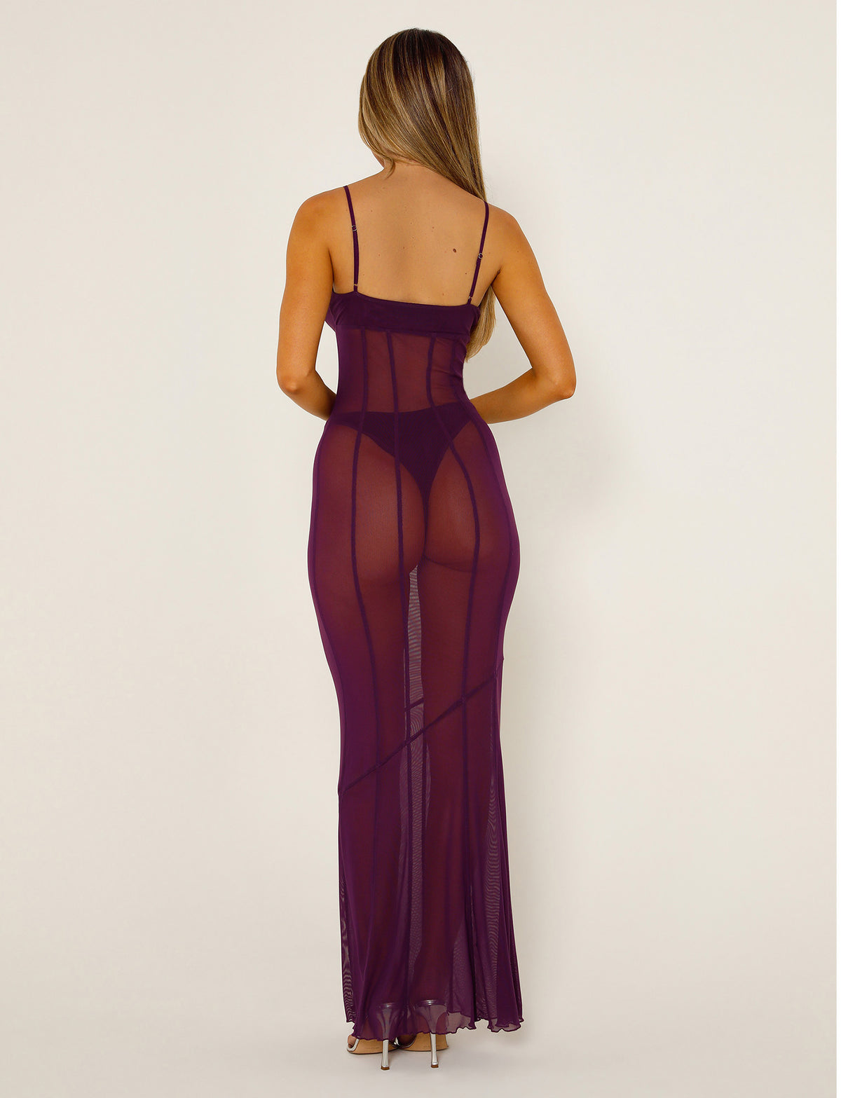 A woman with long hair is facing away from the camera, wearing a deep purple, sheer, form-fitting KAIDA DRESS by TIGERMIST. The floor-length dress has spaghetti straps and she stands against a plain, beige background.