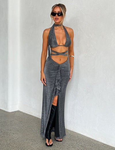 A woman is standing against a white textured wall, showcasing a chic ensemble featuring the TIGERMIST DAISIE SKIRT - BLACK: BLACK SHIMMER. She's accessorized with sunglasses, black heeled boots, and a bracelet on her left arm. Her hair is styled in an elegant updo as she poses confidently looking forward.