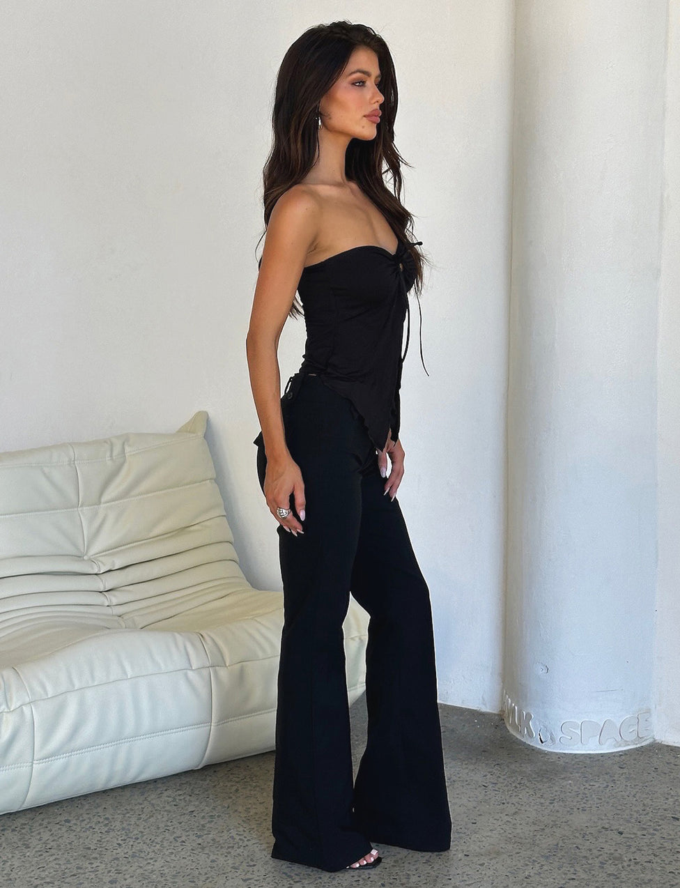 A woman with long, dark hair is standing indoors near a white couch and a white wall. She is wearing the KAROLINA JERSEY TOP in black and black pants by TIGERMIST. She stands in profile, facing to the left, with her hands resting by her sides. The setting appears minimalist and modern.