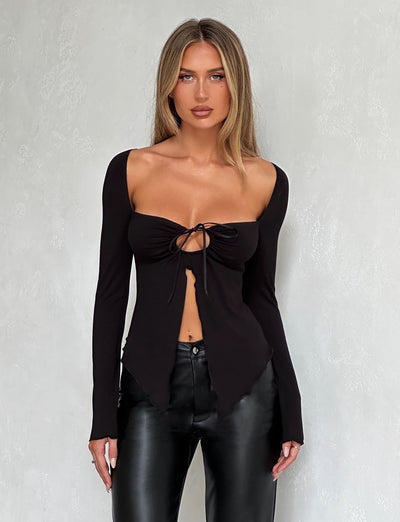 A woman is standing against a light gray textured wall, dressed in TIGERMIST's KAROLINA JERSEY LONG SLEEVE TOP - BLACK and black leather pants. Her blonde hair is styled straight, and she maintains a neutral expression.
