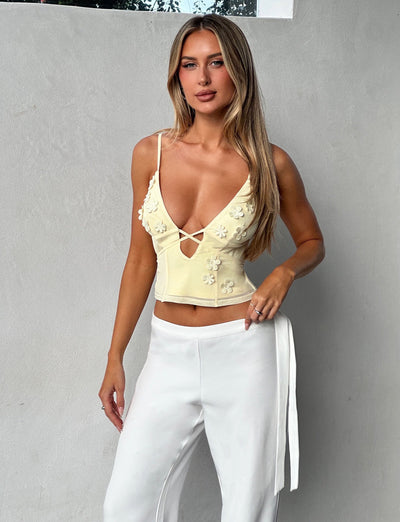A person stands against a light gray wall, wearing the Tiger Mist Katina Cami in Butter yellow, which features flower embellishments and a plunging neckline, paired with white pants with a sash belt. They have long, straight blonde hair and a neutral expression while posing with one hand on their hip.