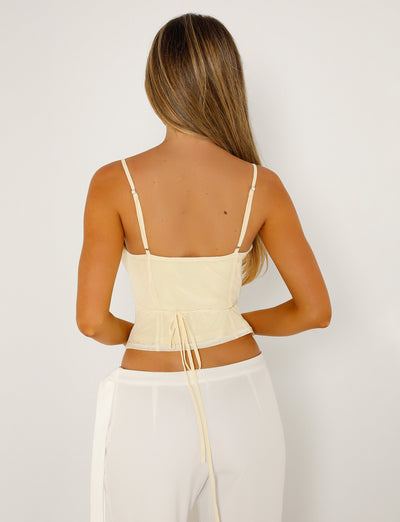 A woman with long, straight hair is shown from the back, wearing the TIGERMIST KATINA CAMI in a light beige color (referred to as "Butter"), a sleeveless, back-tie top with thin straps, and white high-waisted pants. The photo background is plain white.