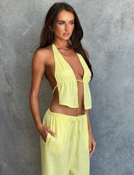 A woman with long dark hair stands in front of a gray textured background, dressed head-to-toe in TIGERMIST. She is wearing the LAINEY TOP - YELLOW : LEMON, a light yellow halter top that ties in the front, and matching light yellow pants with a drawstring waist. With a neutral expression, her hands are casually tucked into her pockets.
