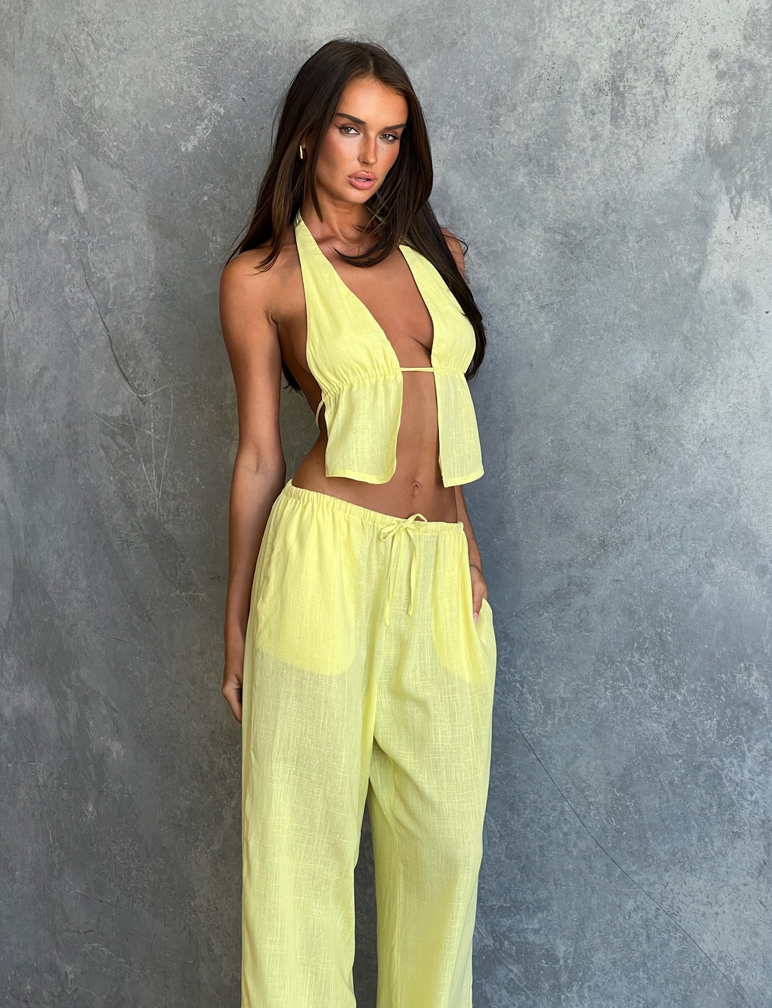A person with long hair, dressed in a yellow halter top and the LAINEY PANT - LEMON by TIGERMIST, poses against a textured gray wall. They have one hand resting on their thigh while looking off-camera with a serious expression.