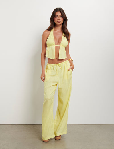 A woman stands against a plain white wall, wearing the LAINEY PANT in Lemon from TIGERMIST. Styled by Tiger Mist, she complements the flowing light yellow halter top and wide-leg pants ensemble with a bracelet. Her long brown hair cascades beautifully as she gazes directly at the camera with a relaxed expression.