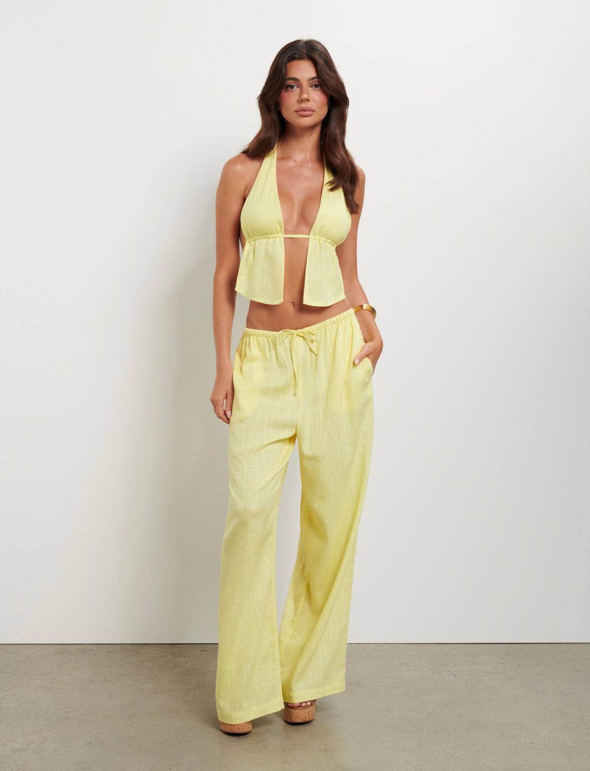 A woman stands against a plain white background, wearing the LAINEY TOP - YELLOW : LEMON from TIGERMIST. The ensemble consists of a halter-style crop top complemented by loose, high-waisted pants. With long, wavy brown hair and beige heels, she gazes directly at the camera.