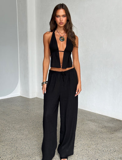 A woman with long brown hair poses against a light gray wall, stunningly dressed in TIGERMIST's black halter top with a deep V-neck and the LAINEY PANT - BLACK. She accessorizes with a large pendant necklace and bracelets on one wrist. The setting appears to be an indoor space with a polished floor.