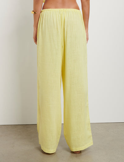 A person is shown from the back, wearing loose-fitting, high-waisted LAINEY PANT - YELLOW : LEMON pants by TIGERMIST. The person is barefoot and standing on a smooth, light-colored surface in front of a plain white wall.