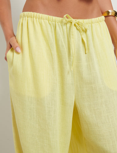 The image shows a close-up of a person wearing the LAINEY PANT - YELLOW: LEMON by TIGERMIST. These high-waisted, loose-fitting pants feature an elastic waistband with a drawstring. The person's hands are tucked into the front pockets, and a golden bracelet is visible on their right wrist.