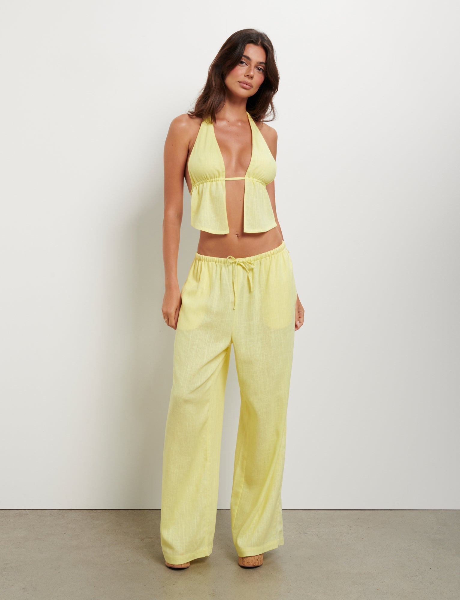 A woman wearing a TIGERMIST LAINEY TOP - YELLOW : LEMON, coupled with matching loose, wide-legged pants, stands against a plain white wall. She poses with her left hand resting on her hip and her right hand relaxed by her side.