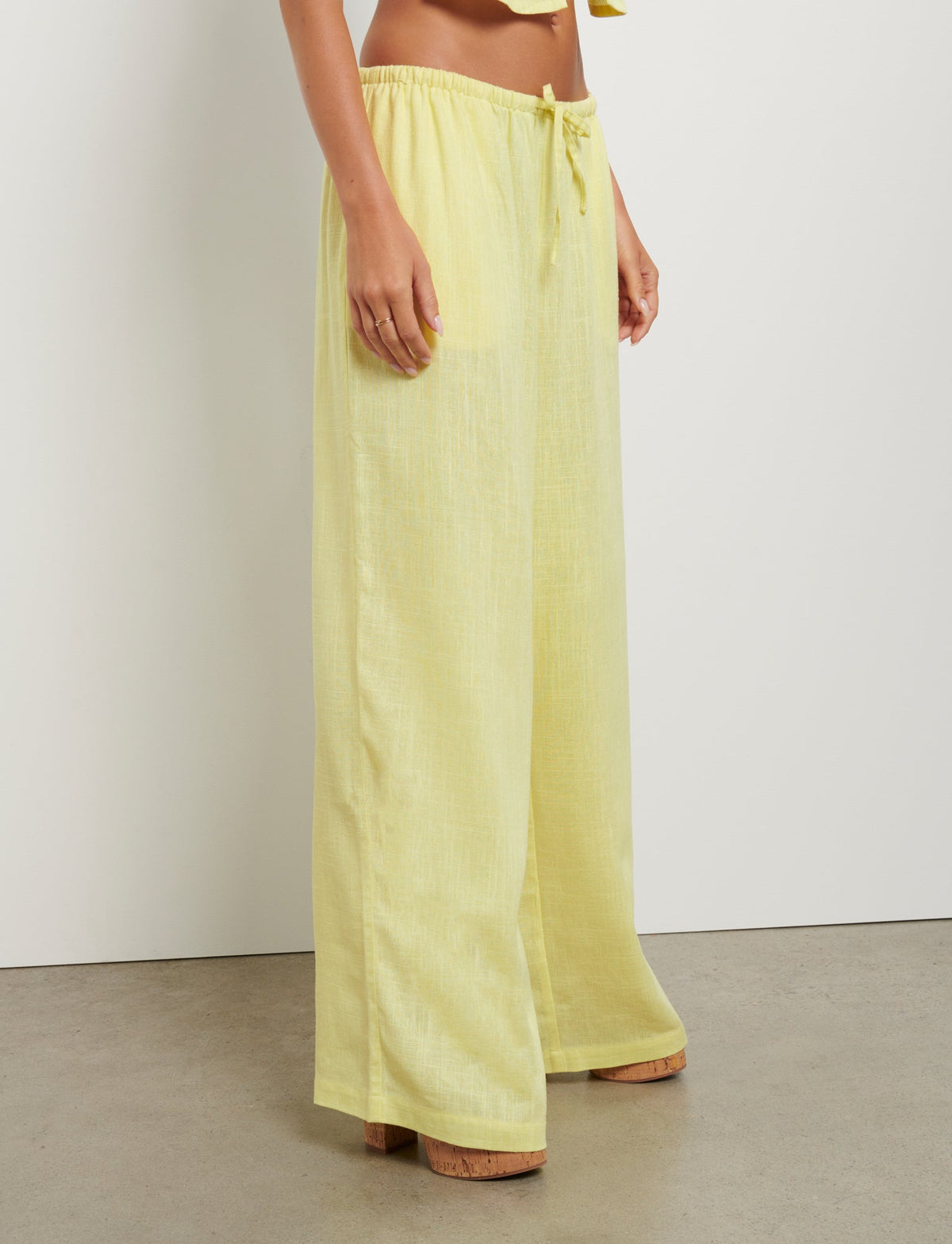 A person wearing the LAINEY PANT - YELLOW : LEMON from TIGERMIST, which are loose, ankle-length trousers with a drawstring waist, stands on a concrete floor against a white wall. The individual is mid-frame with the upper body out of the frame, showing only the hands and lower body.
