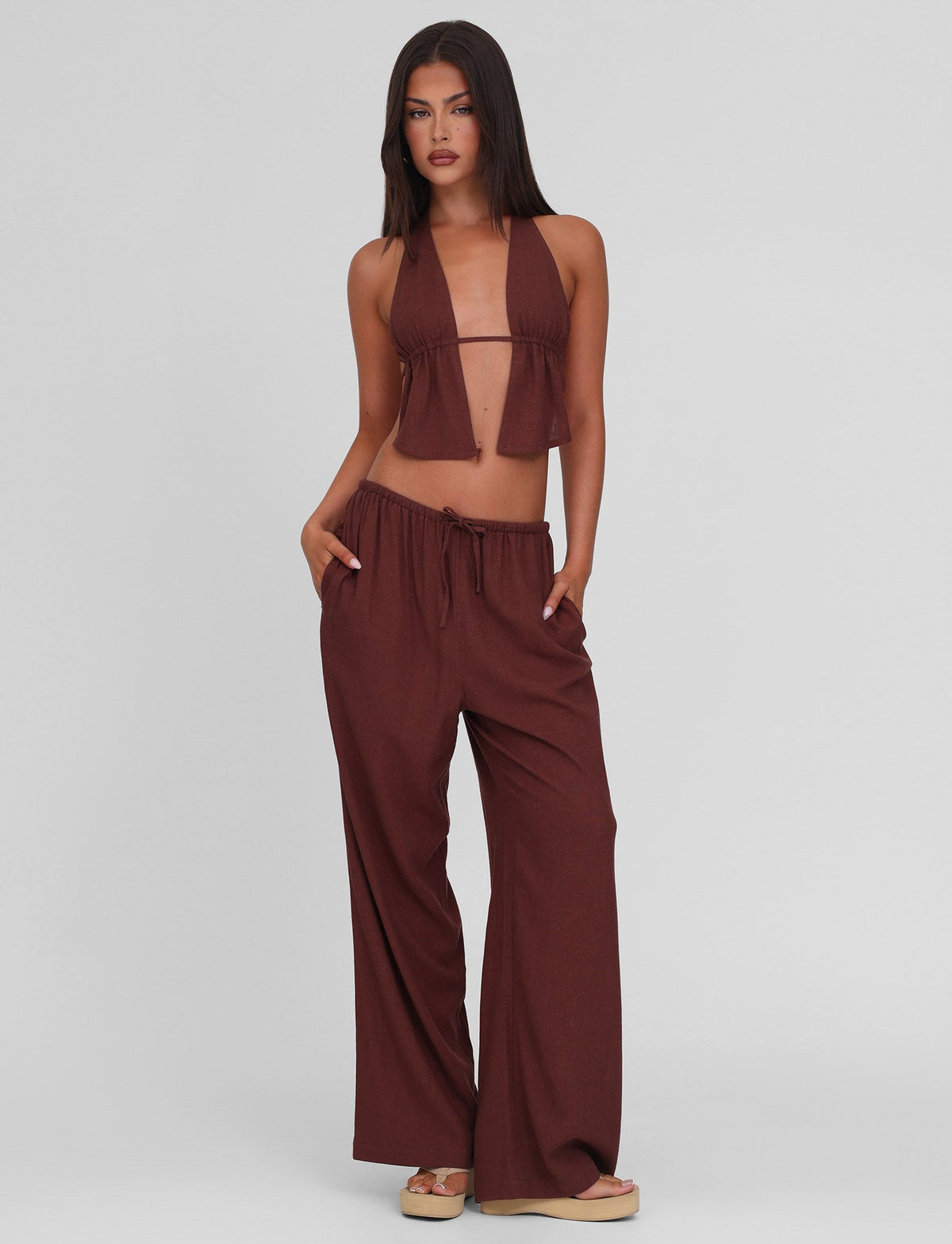 A woman with long dark hair stands against a plain background wearing the TIGERMIST LAINEY PANT in Chocolate—a mid-rise, wide leg linen pant with an elasticated waist—paired with a brown halter crop top and sandals, hands in her pockets.