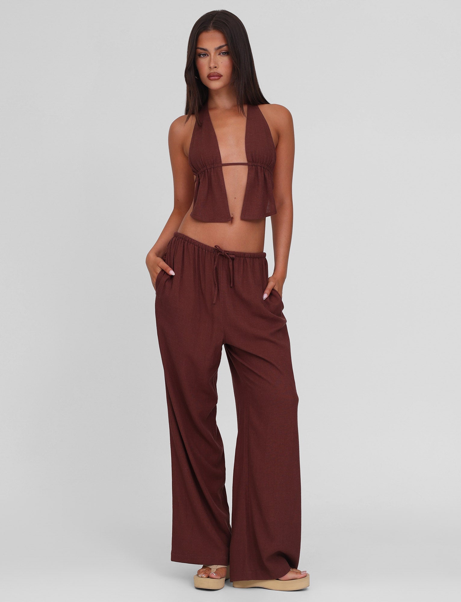 A woman with long dark hair stands against a plain background wearing the TIGERMIST LAINEY PANT in Chocolate—a mid-rise, wide leg linen pant with an elasticated waist—paired with a brown halter crop top and sandals, hands in her pockets.