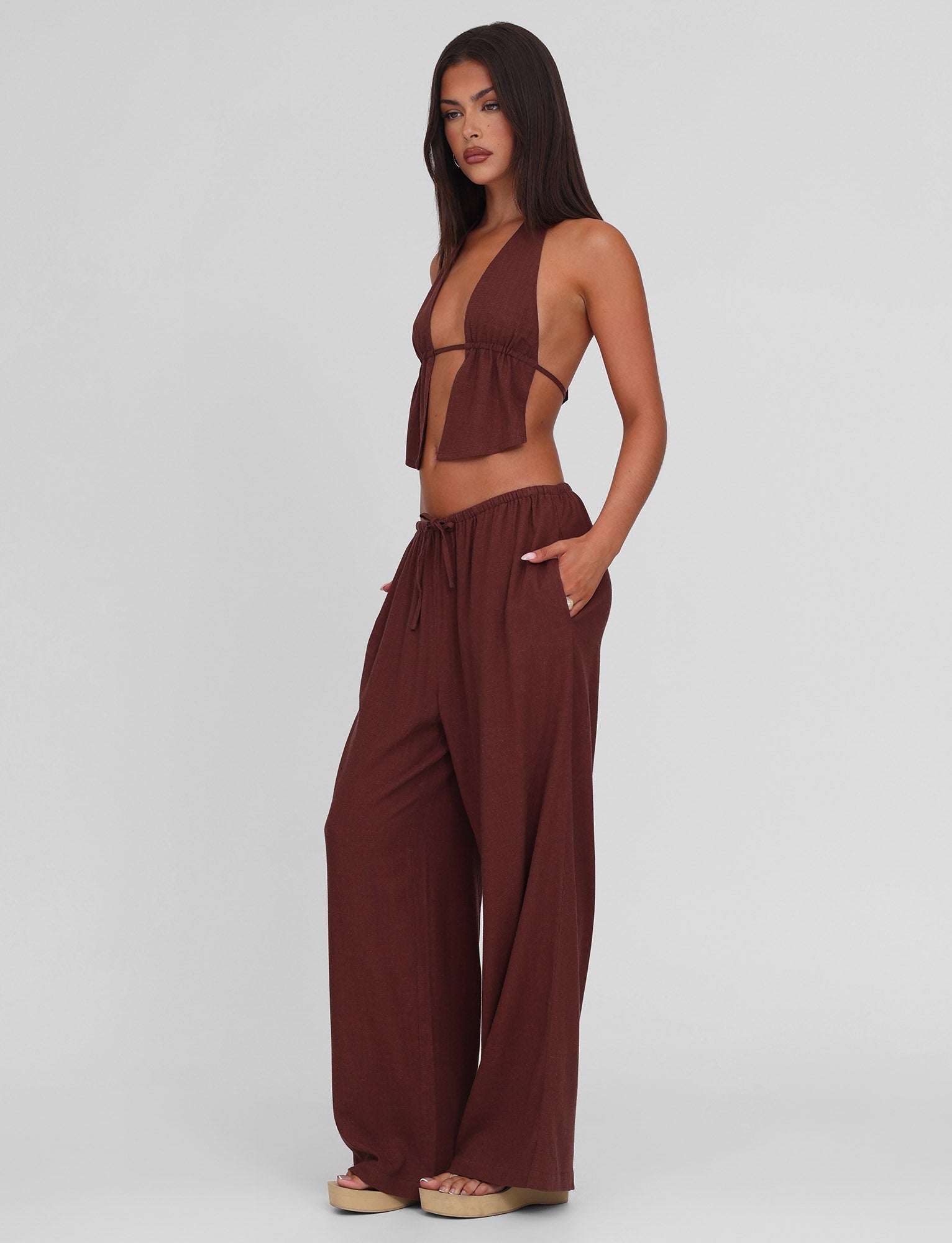 A woman with long dark hair stands against a plain background, wearing TIGERMIST's LAINEY PANT in chocolate—a wide leg linen pant with an elasticated waist—paired with a sleeveless halter top and beige platform sandals. One hand rests in her pocket.