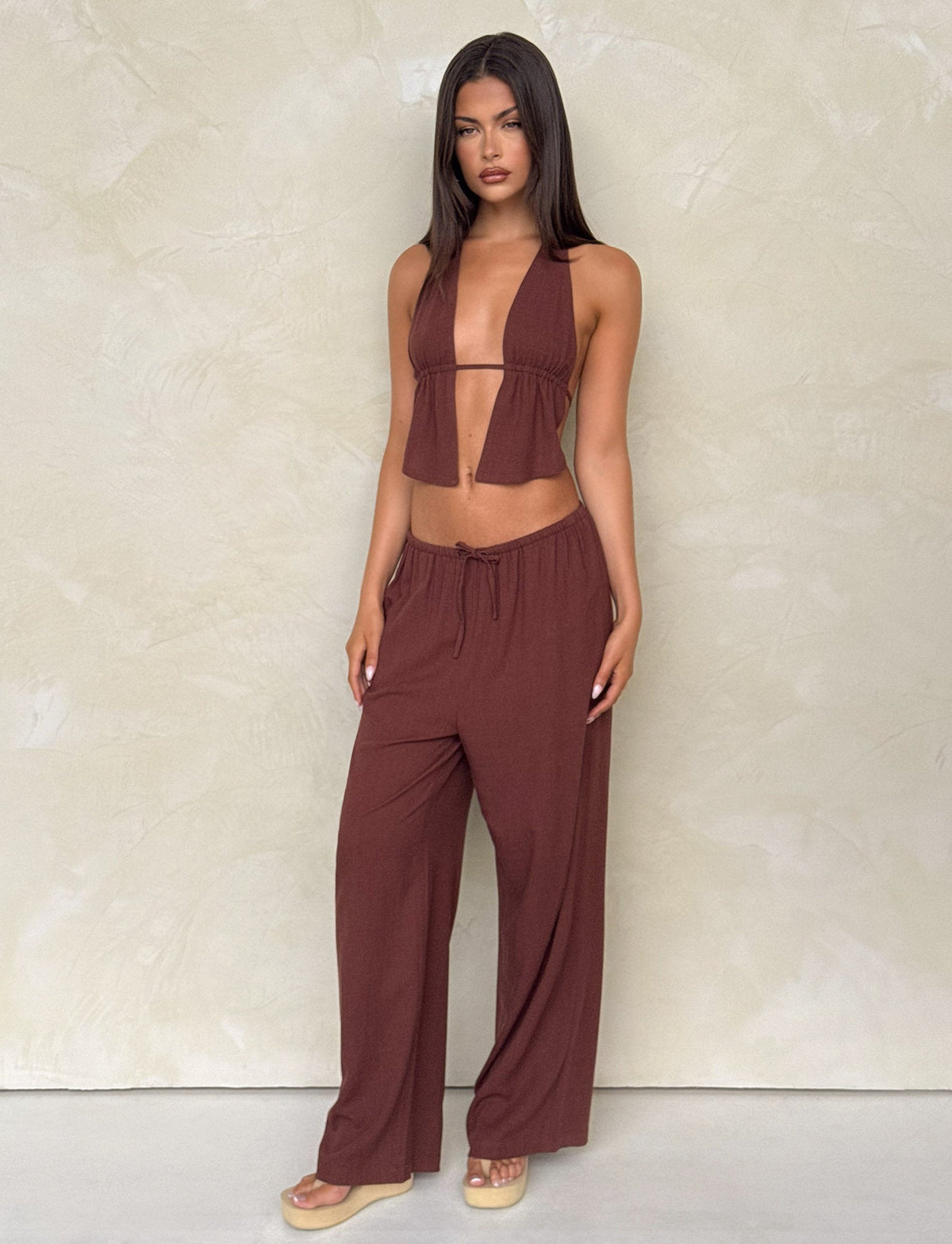 A woman stands against a light textured wall, wearing TIGERMIST's LAINEY PANT in chocolate with an elasticated waist, a matching brown halter crop top, sandals, and has long straight dark hair.