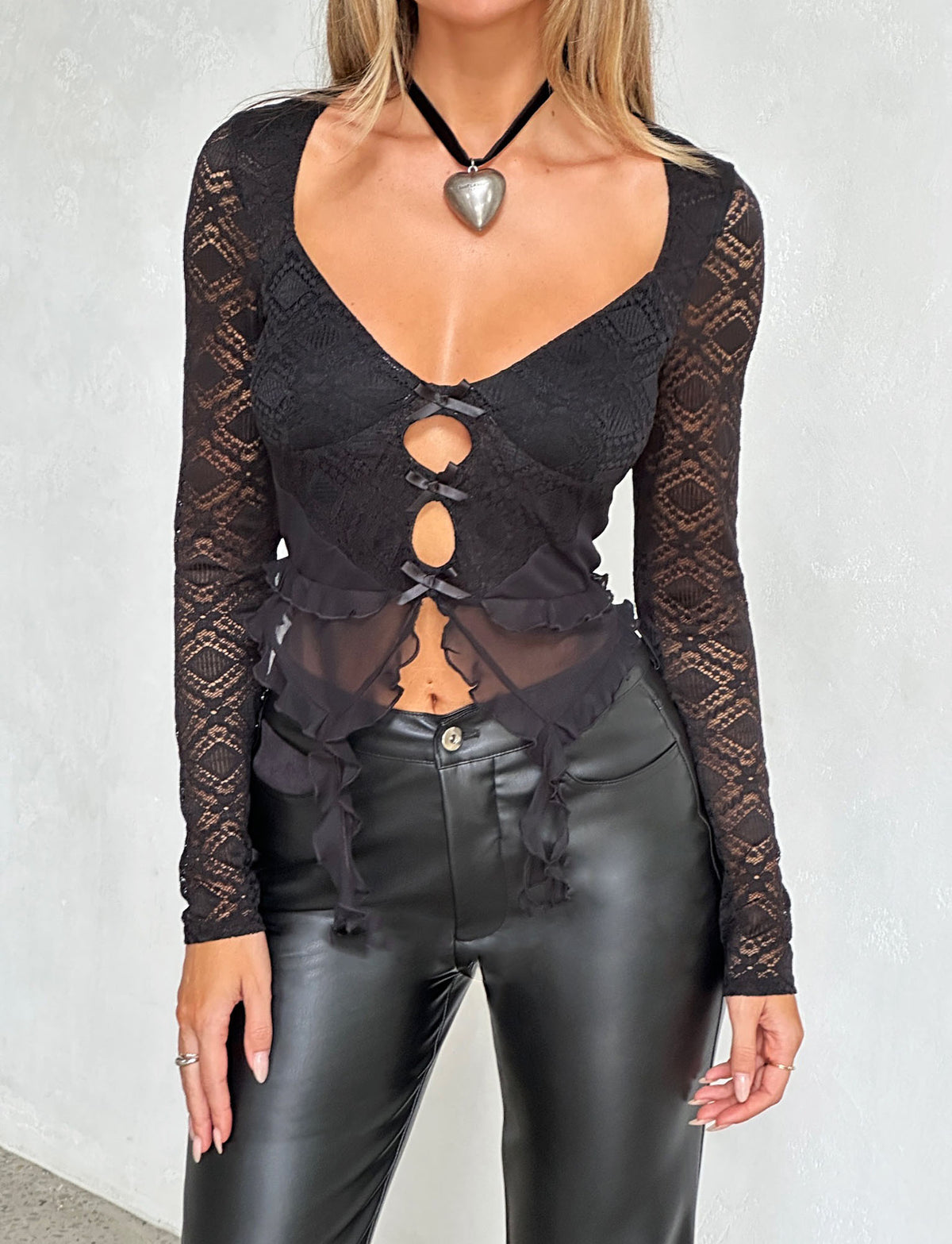 A person is wearing the LAFAYETTE LONG SLEEVE TOP - BLACK by TIGERMIST, featuring long sleeves and three front cut-outs tied with bows. The top includes a heart-shaped pendant attached to a choker. They are also wearing fitted black leather pants against the backdrop of a plain white wall.