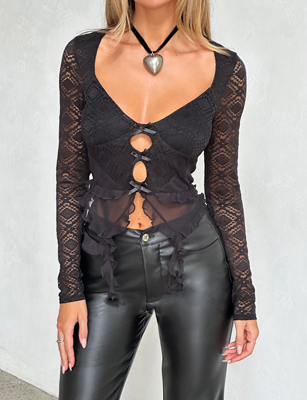 A person is wearing the LAFAYETTE LONG SLEEVE TOP - BLACK by TIGERMIST, featuring long sleeves and three front cut-outs tied with bows. The top includes a heart-shaped pendant attached to a choker. They are also wearing fitted black leather pants against the backdrop of a plain white wall.