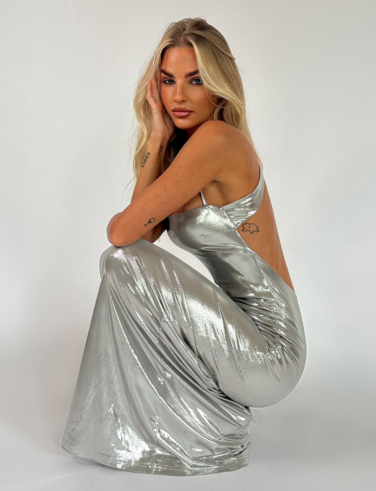 A woman with long blonde hair is crouching down, wearing the ELVIA MAXI DRESS in grey/silver metallic from TIGERMIST. Her hands are resting on her face and knee, and she has tattoos on her forearm. The background is plain white.