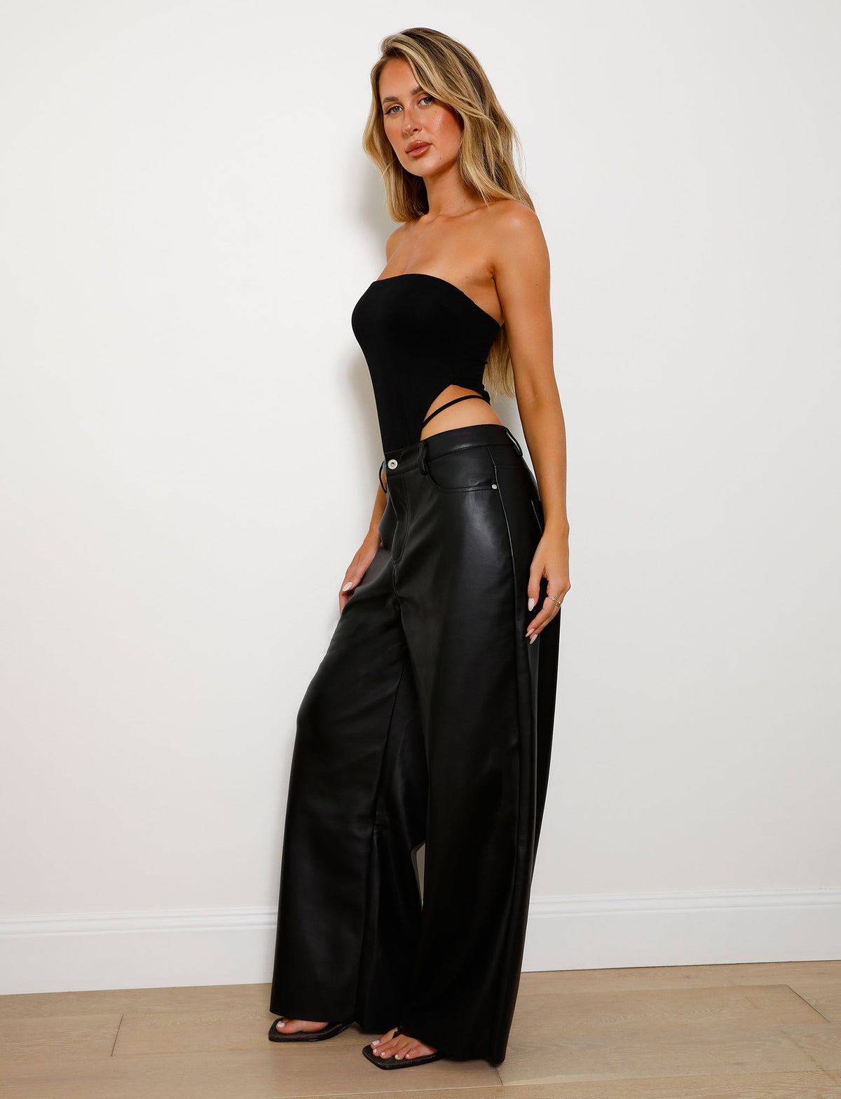 A woman stands against a plain white wall wearing the FINIAN BODYSUIT in black from TIGERMIST, featuring side cutouts. She pairs it with high-waisted, wide-leg black leather pants. With her long, wavy blonde hair cascading down, she poses confidently with one hand on her hip, looking directly at the camera.