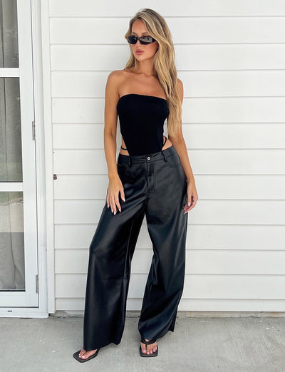 A person with long blonde hair is posing against a white-paneled wall, showcasing a chic outfit from TIGERMIST. They are wearing a black strapless top, the LELA PANT in black, black flip-flops, and black sunglasses.