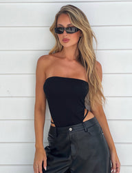 A woman with long blonde hair is wearing the FINIAN BODYSUIT - BLACK from TIGERMIST, paired with black leather pants and black sunglasses. She stands against a white background with slight grooves, her left hand resting on her hip and a relaxed expression on her face.