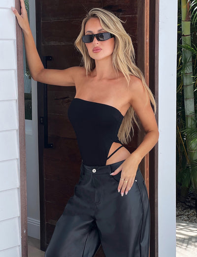A woman with long blonde hair, wearing black sunglasses and a Tiger Mist strapless black bodysuit with side cutouts, pairs it stylishly with the TIGERMIST LELA PANT in black. She leans against a wooden door, one hand on the door frame and the other resting in her pocket.