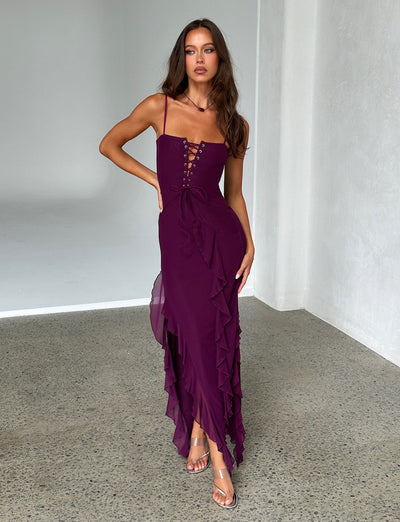A woman stands in a minimalistic room wearing the deep purple CHAYA DRESS from TIGERMIST. The sleeveless dress features lace-up detailing and ruffles along the side. She poses with one hand on her hip and the other relaxed while gazing confidently forward, complemented by her transparent high-heeled sandals.