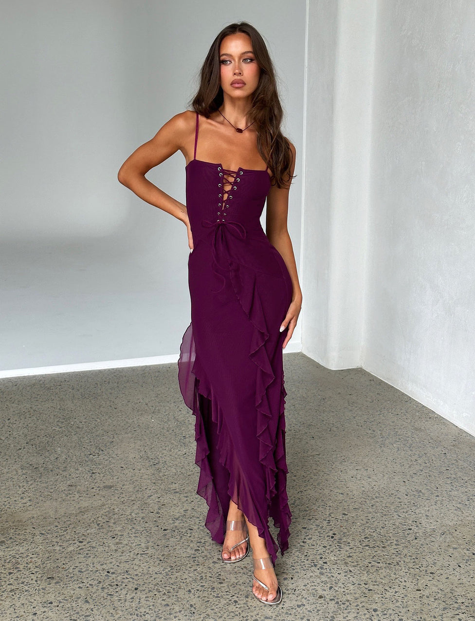 A woman stands in a minimalistic room wearing the deep purple CHAYA DRESS from TIGERMIST. The sleeveless dress features lace-up detailing and ruffles along the side. She poses with one hand on her hip and the other relaxed while gazing confidently forward, complemented by her transparent high-heeled sandals.
