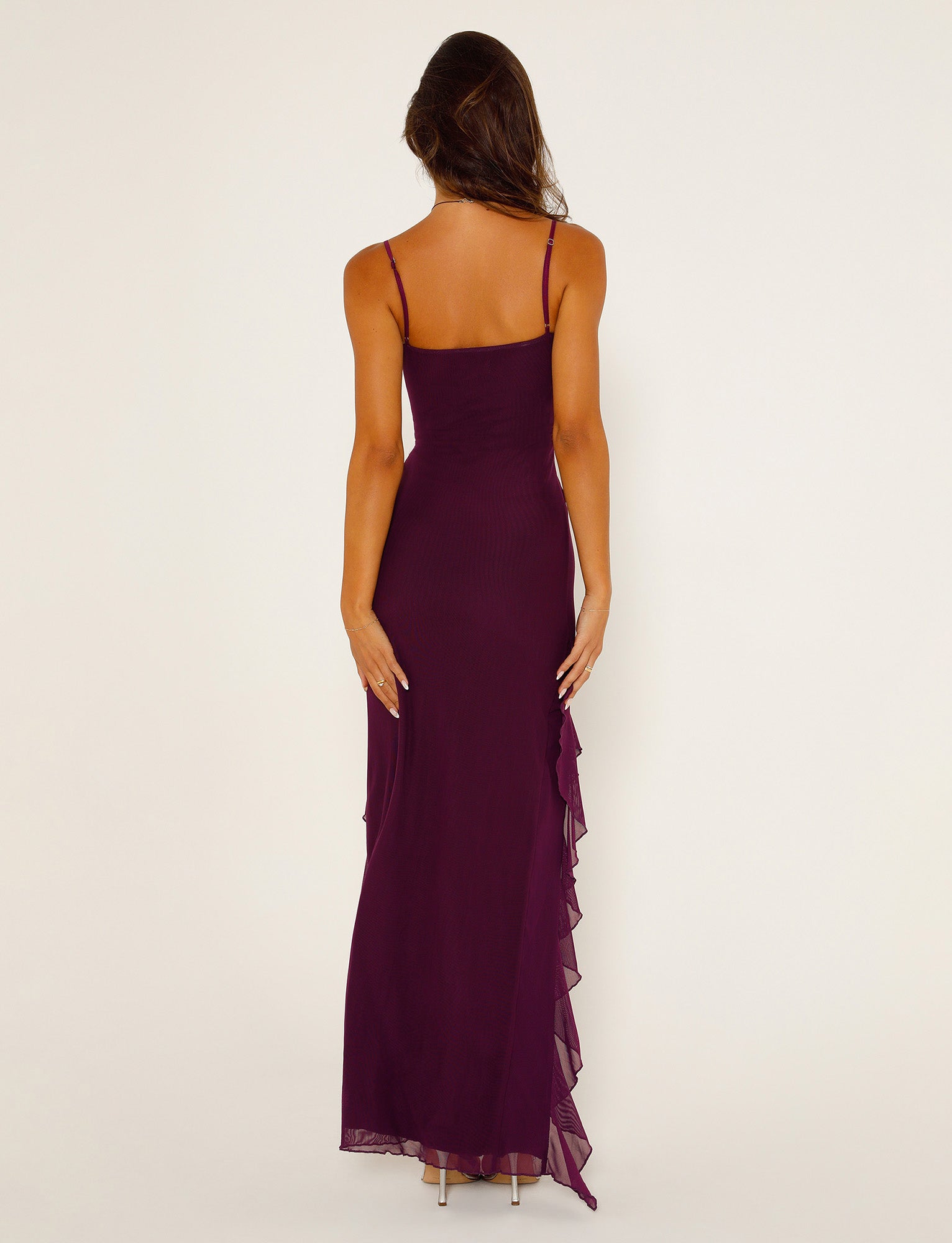 A woman with long, wavy hair is standing with her back to the camera, wearing a TIGERMIST CHAYA DRESS in deep purple. The sleeveless floor-length gown features thin straps and a layered ruffle detail on the side. She is also wearing high-heeled shoes.