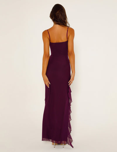 A woman with long, wavy hair is standing with her back to the camera, wearing a TIGERMIST CHAYA DRESS in deep purple. The sleeveless floor-length gown features thin straps and a layered ruffle detail on the side. She is also wearing high-heeled shoes.