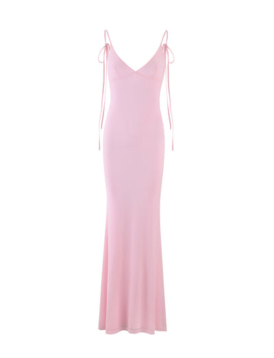 The KYLAH DRESS - PINK : BABY PINK from TIGERMIST is a full-length pink dress with a fitted silhouette, featuring thin spaghetti straps and a deep V-neckline. It includes additional ties that drape off the shoulders, adding a delicate touch. Its smooth, flowing fabric flares slightly at the hem.