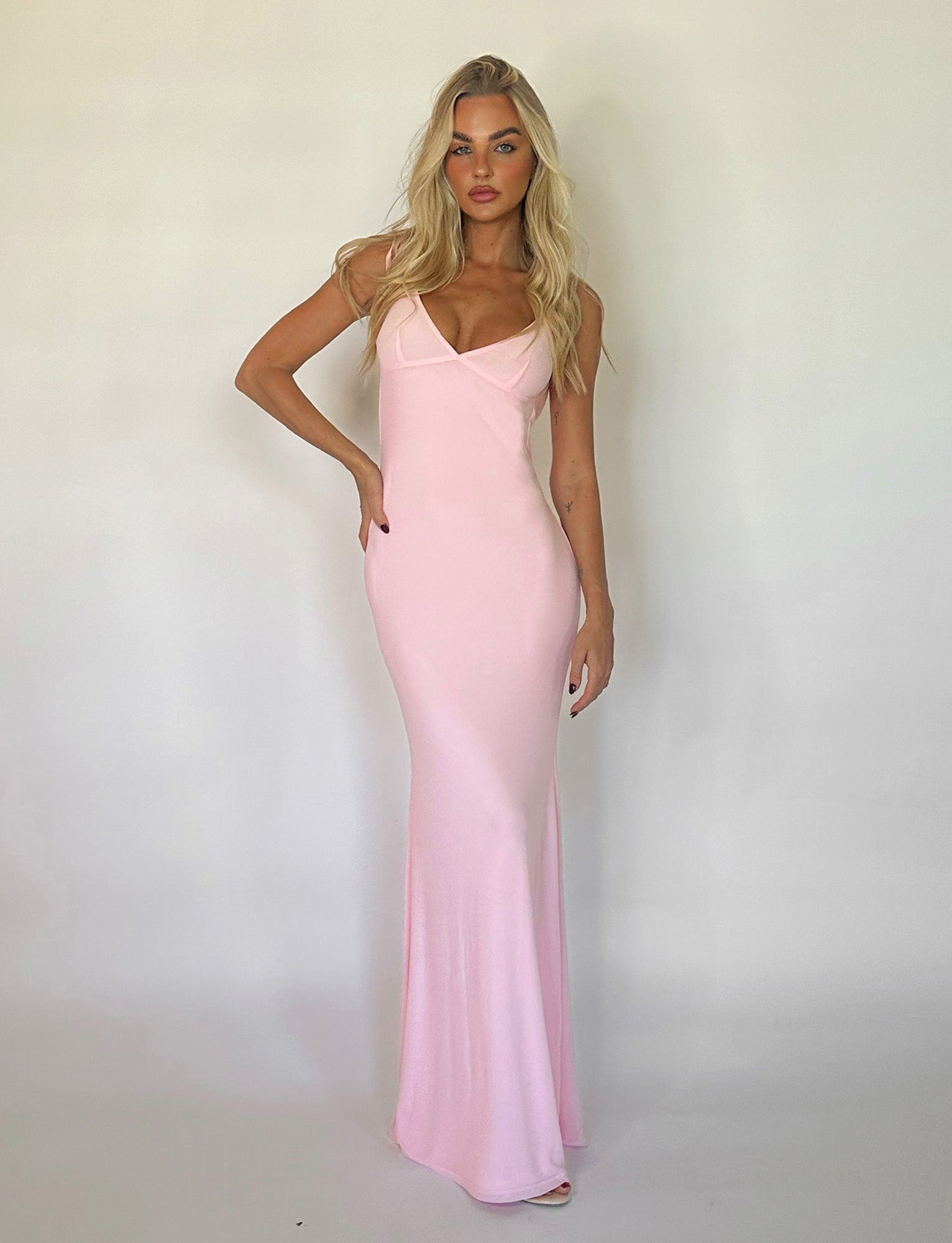A woman with long blonde hair stands against a plain white background, wearing a floor-length, sleeveless KYLAH DRESS in baby pink by TIGERMIST. She is posing with one hand on her hip and the other hanging loosely by her side.