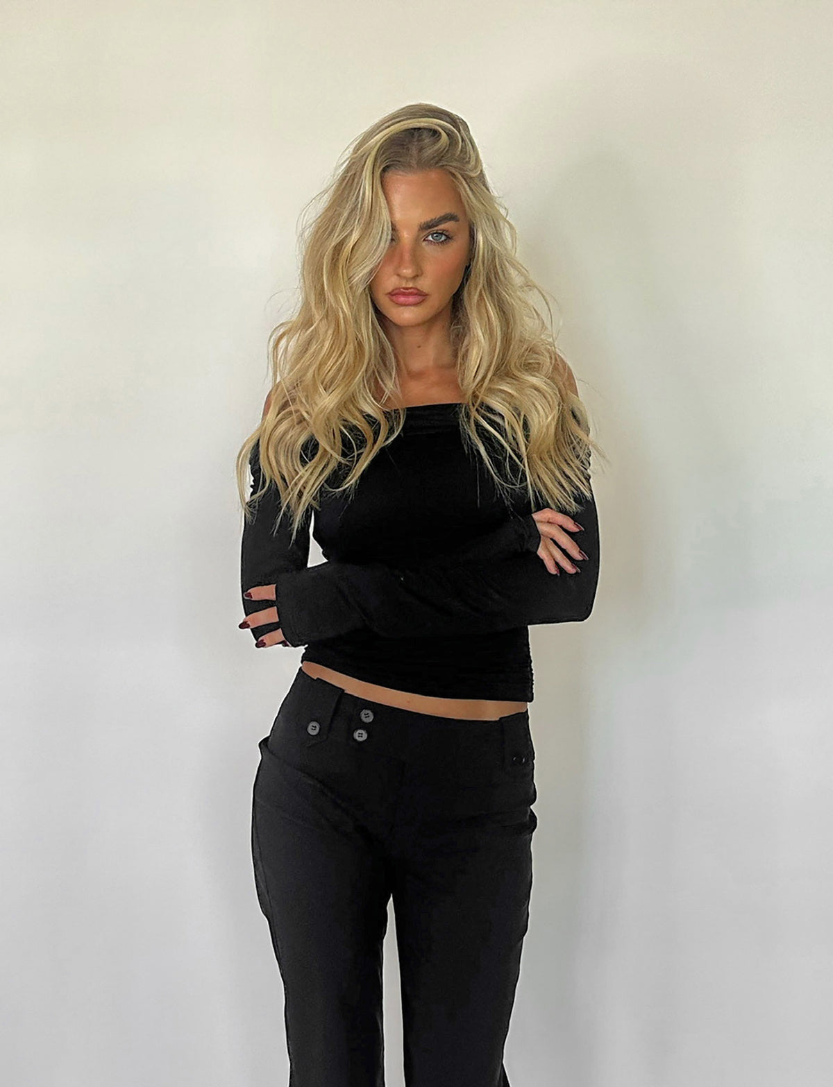 A person with long, wavy blonde hair stands against a plain white background. They are wearing the INDIANA TOP - BLACK by TIGERMIST, featuring an off-the-shoulder design and long sleeves, paired with matching black pants. Their arms are crossed in front of them, and they have a serious expression.
