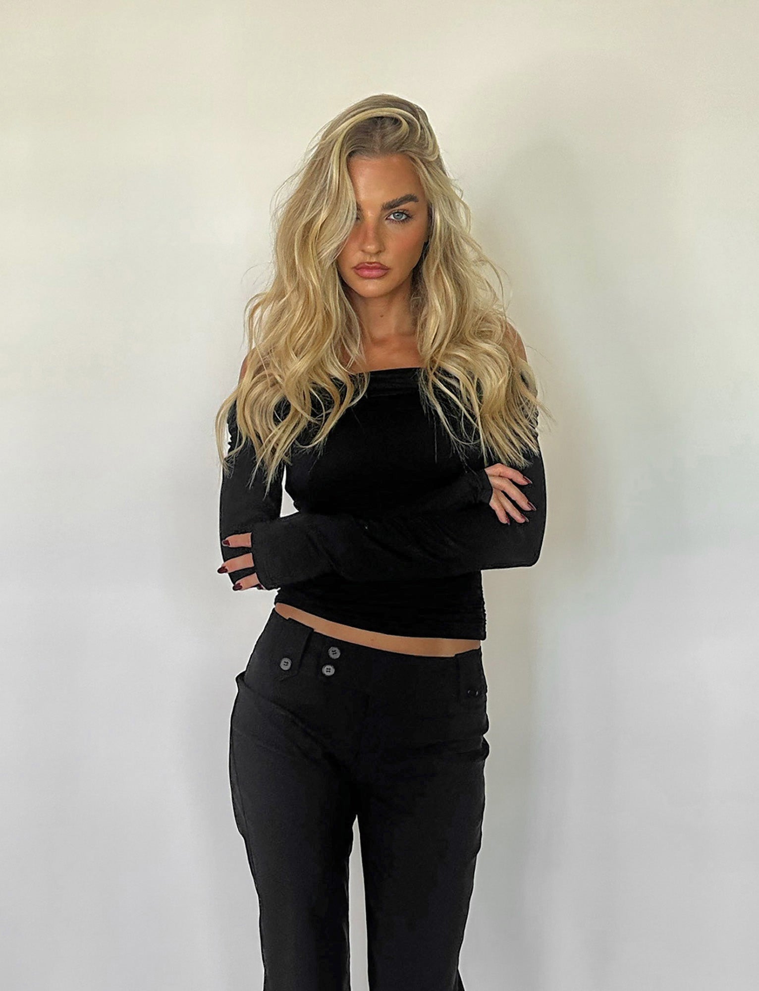 A person with long, wavy blonde hair stands against a plain white background. They are wearing the INDIANA TOP - BLACK by TIGERMIST, featuring an off-the-shoulder design and long sleeves, paired with matching black pants. Their arms are crossed in front of them, and they have a serious expression.