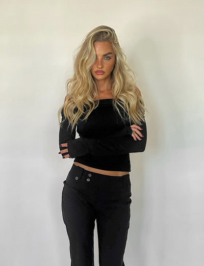 A person with long, wavy blonde hair stands against a plain white background. They are wearing the INDIANA TOP - BLACK by TIGERMIST, featuring an off-the-shoulder design and long sleeves, paired with matching black pants. Their arms are crossed in front of them, and they have a serious expression.