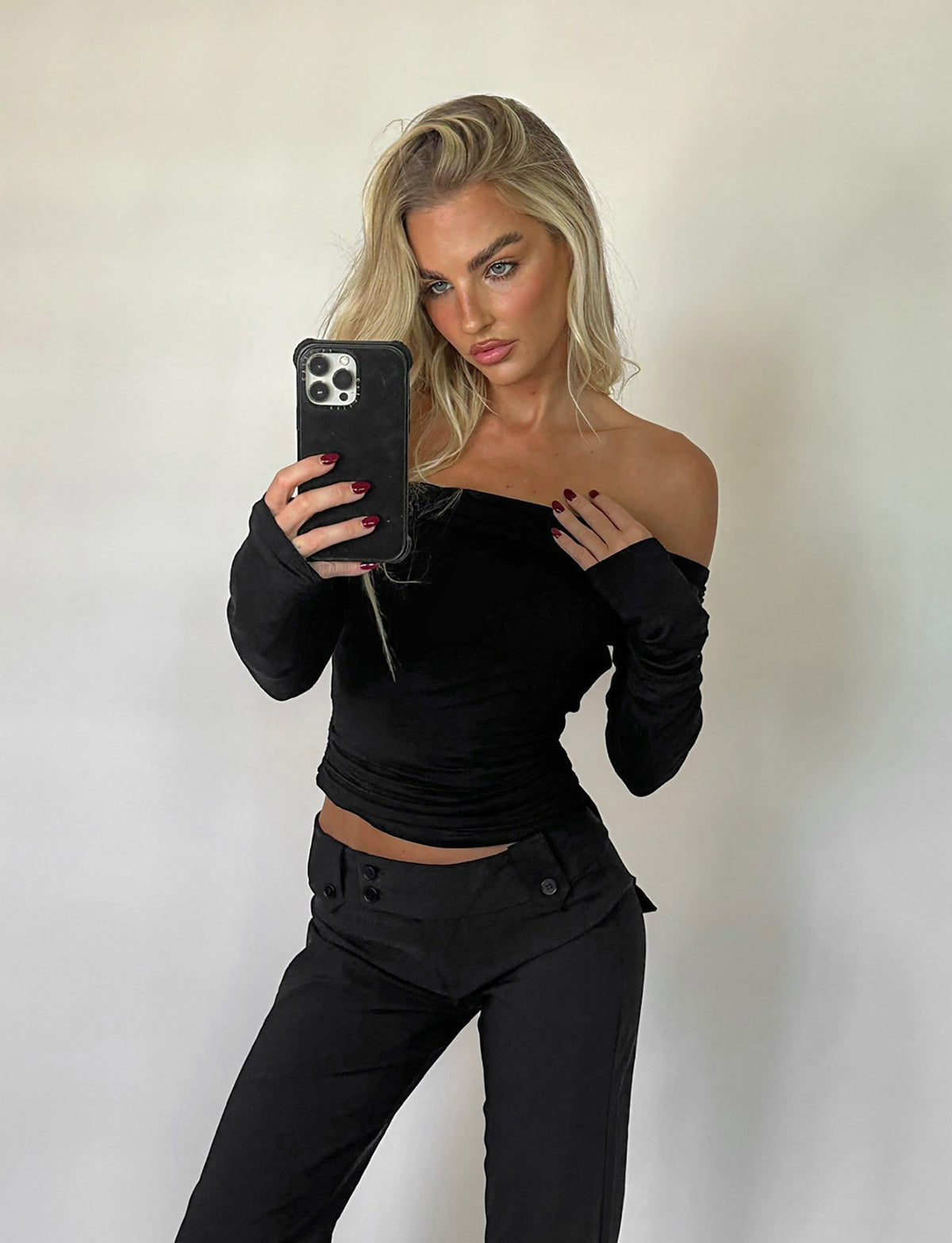 A woman with long blonde hair takes a mirror selfie with her phone. She is dressed in the INDIANA TOP - BLACK from TIGERMIST and matching black pants. The background is plain and light-colored.