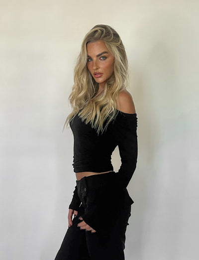 A woman with long, wavy blonde hair stands against a plain white background. She is wearing the chic INDIANA TOP in black and matching black pants from TIGERMIST. Her expression is neutral, and her hands rest at her sides.